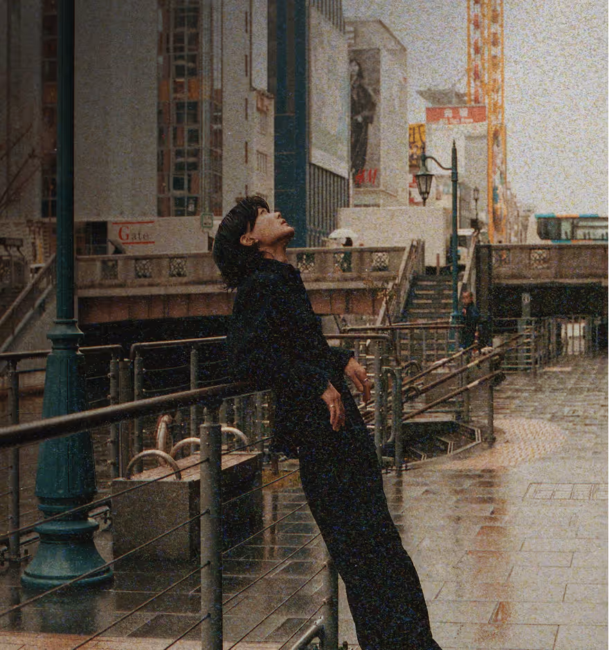Grainy image of a person dressed in black leaning back against a railing on a rainy city sidewalk with tall buildings in the background.