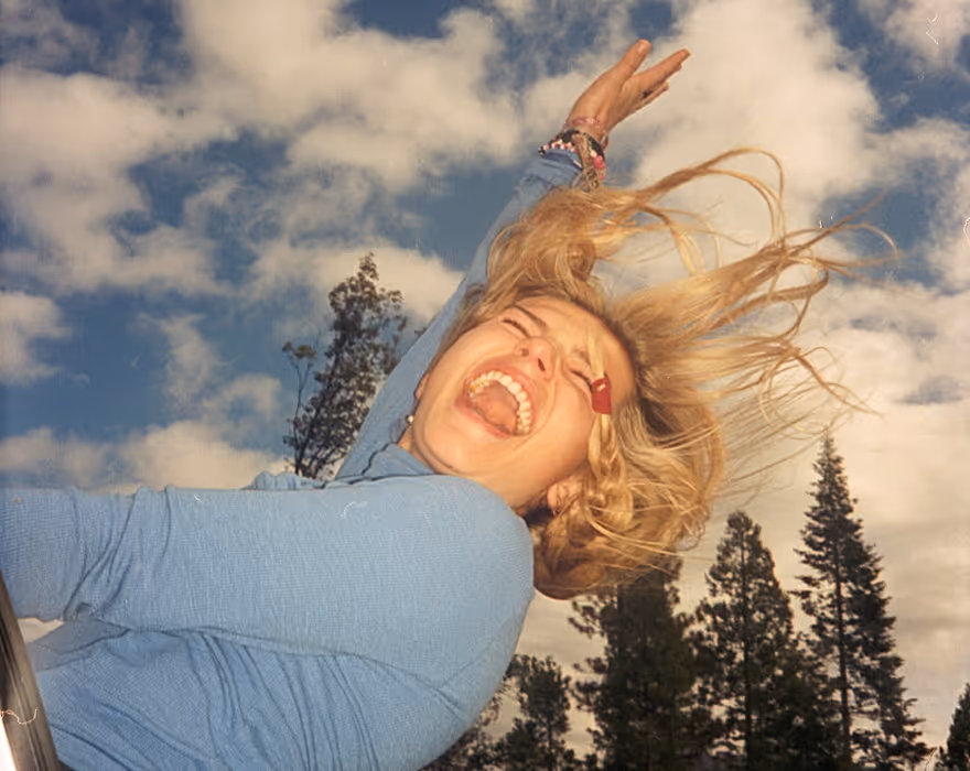 Woman with wind in her blonde hair laughing joyfully leaning out of a moving car window against a backdrop of blue sky with clouds and tall pine trees.