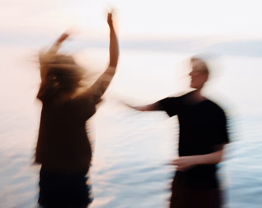 Motion blurred video still of two people by water, one with arms raised and flowing hair, the other facing them.