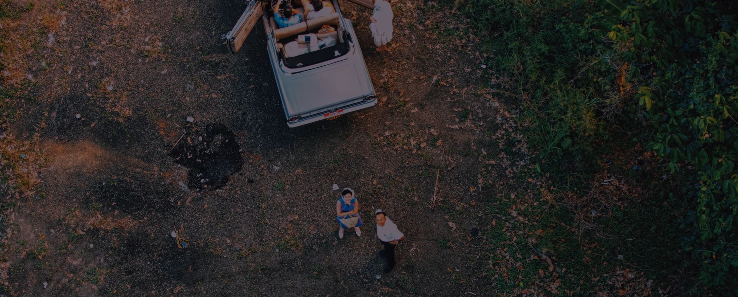 Aerial view of three people near a silver convertible car parked on a gravel path beside green foliage.
