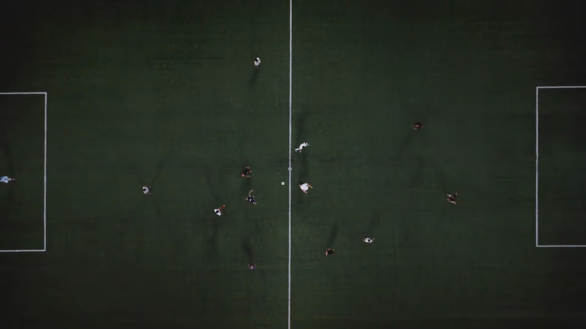 Video still of an aerial view of soccer players on a green field near the center line and ball.