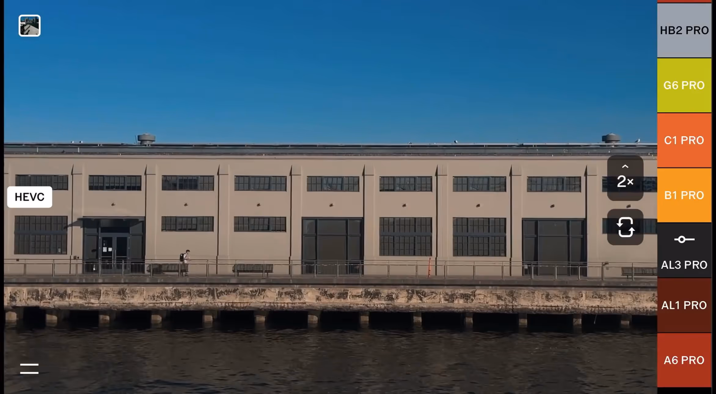 Horizontal video recording in the VSCO Capture camera app with live camera filters menu on the righthand side, showing a waterfront view of a beige industrial building with multiple large windows under a clear blue sky.