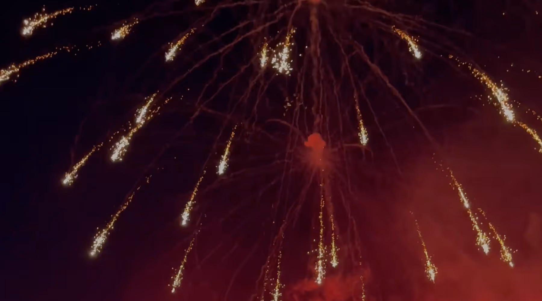 Golden fireworks streaks falling against a dark night sky with red smoke below.