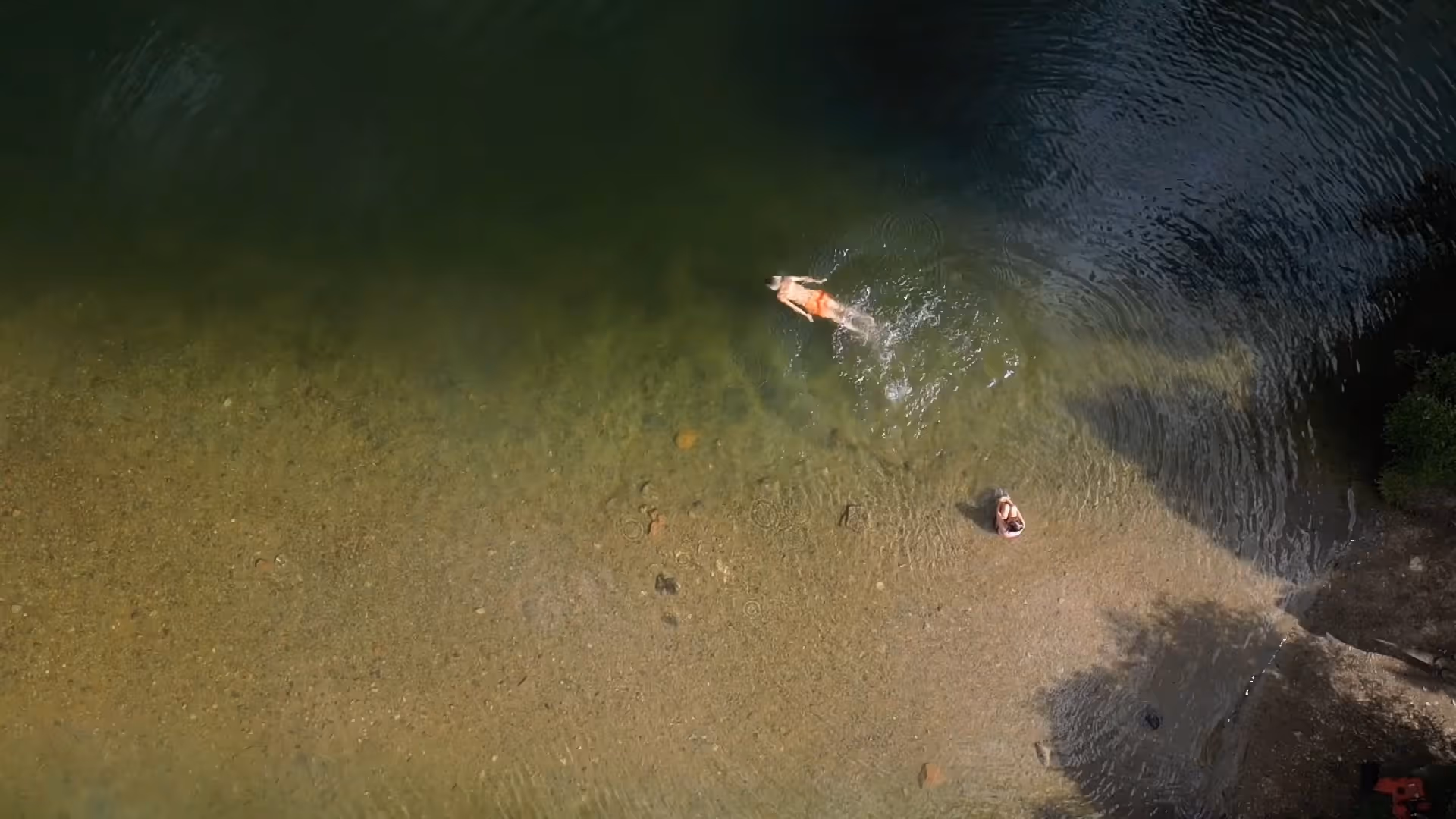 Slow motion video aerial view of a person swimming in clear shallow water near a sandy, pebbly shore where another person sits.