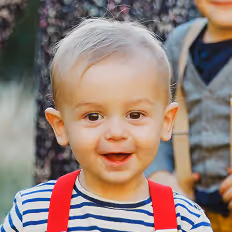 Batch edited family photoshoot of a smiling toddler with light hair wearing a striped shirt and red suspenders outdoors.