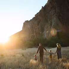 Batch edited family photoshoot of a family of four holding hands and walking through tall grass with sun setting behind rocky cliffs.