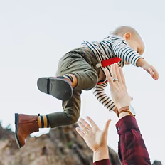 Batch edited family photoshoot of a child in striped shirt and boots being lifted into the air by adult hands outdoors.