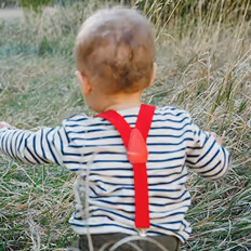 Batch edited family photoshoot of a baby boy with red suspenders on striped shirt sitting in tall grass outdoors.