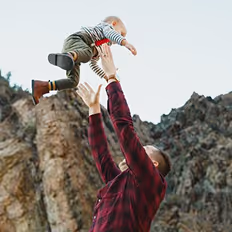 Batch edited family photoshoot of a man lifting child high in the air outdoors with rocky hills in the background.
