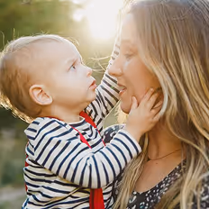 Batch edited family photoshoot of a baby in striped shirt touching a woman's face outdoors with sunlight behind them.