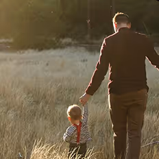 Batch edited family photoshoot of a father and child holding hands walking through tall grass in a sunlit outdoor setting.