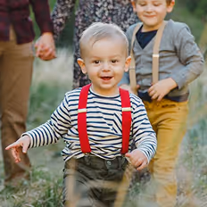 Batch edited family photoshoot of a smiling toddler in striped shirt and red suspenders walking outdoors with family in background.