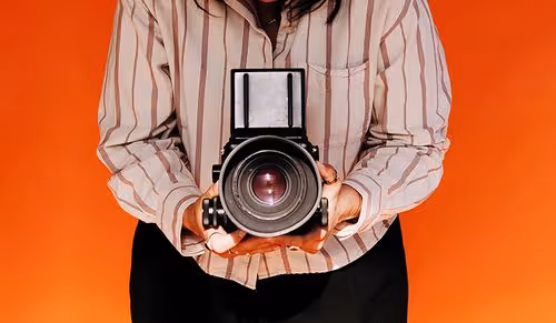 Photographer in striped shirt holding a large camera, looking down at it against orange background.
