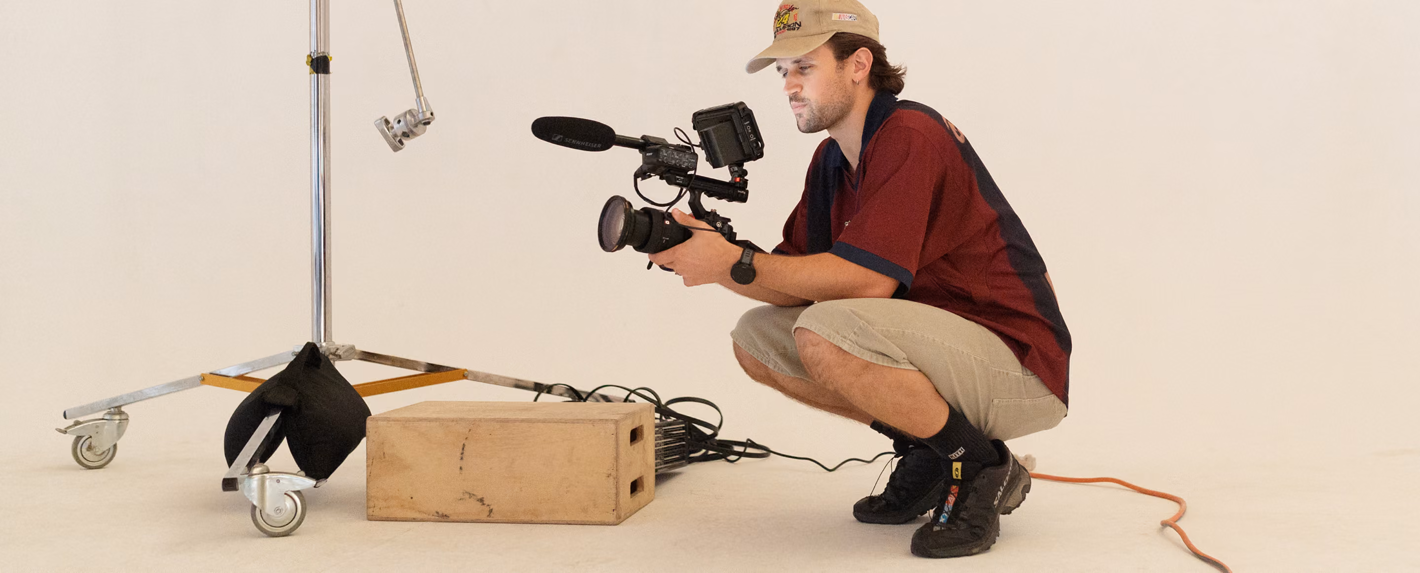 Photographer squatting with video camera in a studio next to a wooden box and light stand on wheels.