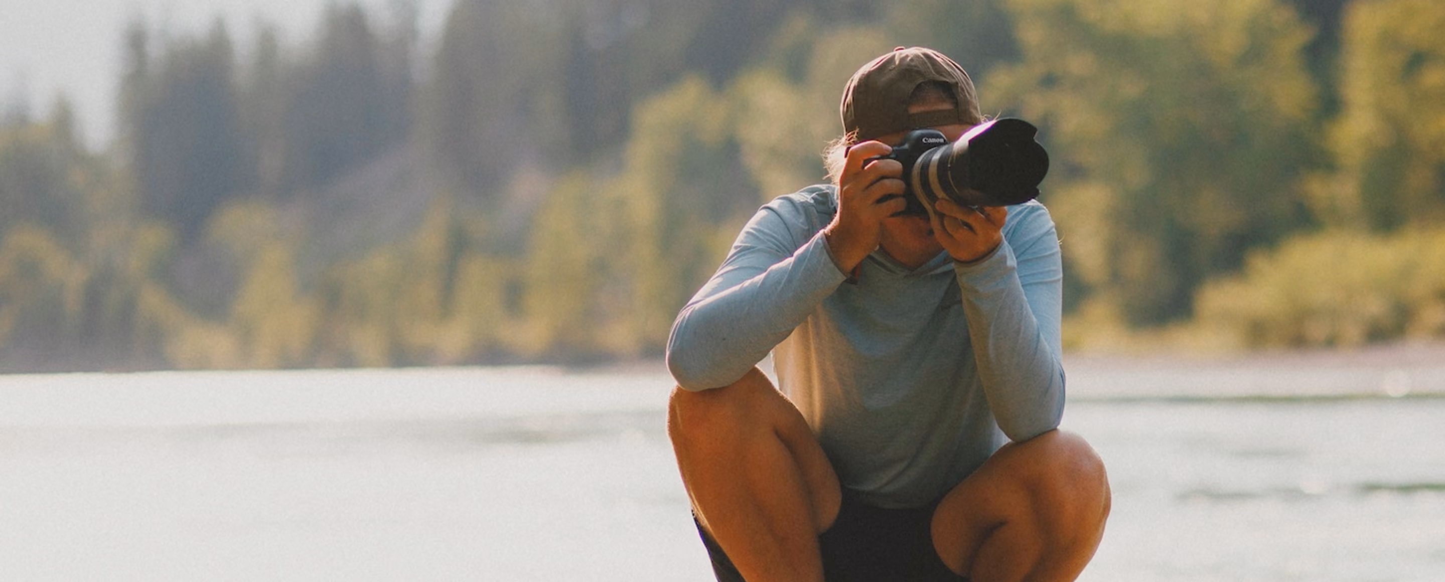 Person squatting outdoors by water, taking a photo with a Canon camera and wearing a cap and long sleeve shirt.