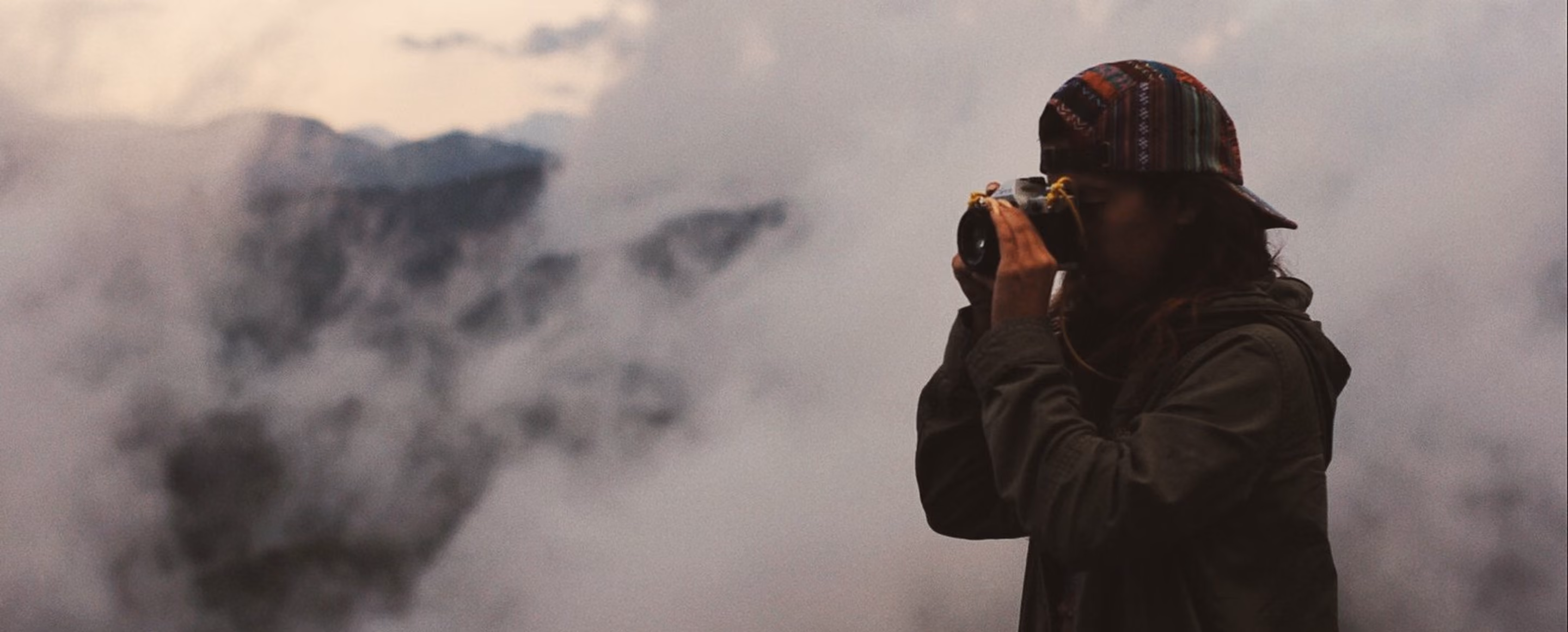 Person in a jacket and striped cap taking a photo with a camera in a foggy mountainous area.