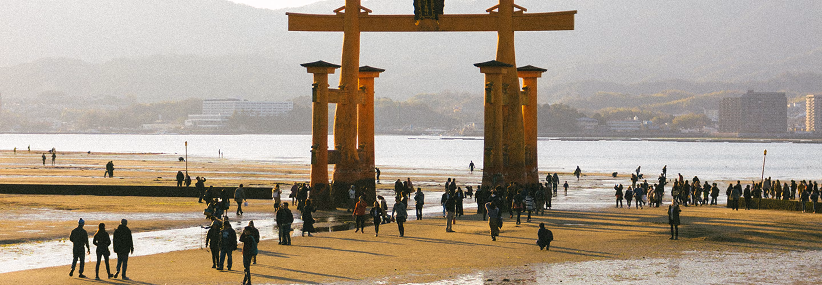 People walking around the large red torii gate at low tide on a beach with buildings in the background.