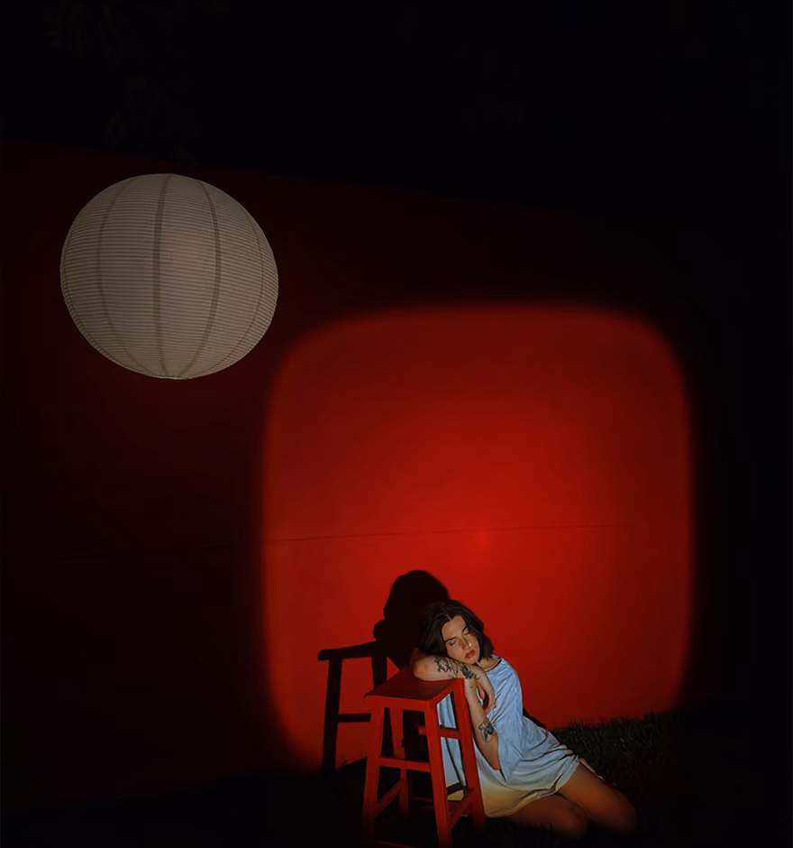 Woman in white shirt leans on red stool in front of red-lit wall under a white lantern at night.