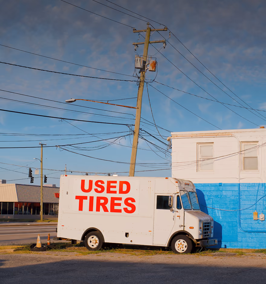 White truck with large red 'USED TIRES' text parked by a building with a blue wall under a clear sky.