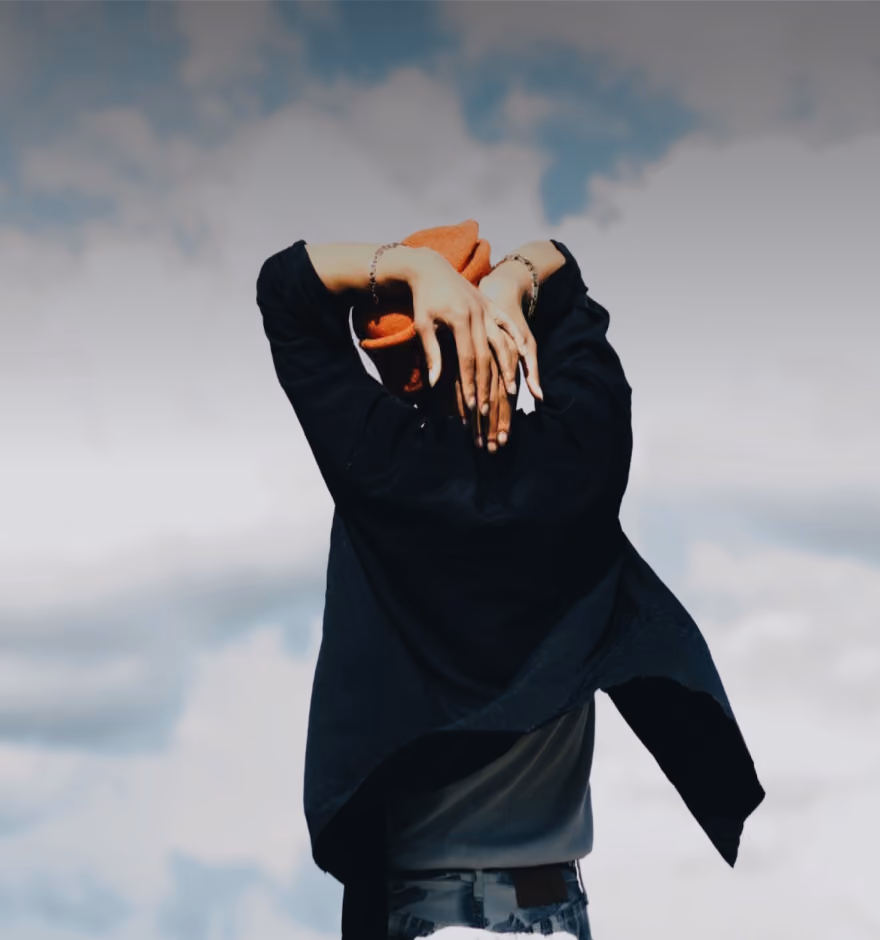 Person with back to camera wearing an orange beanie, black jacket, and bracelets, raising hands to head against a cloudy sky background.