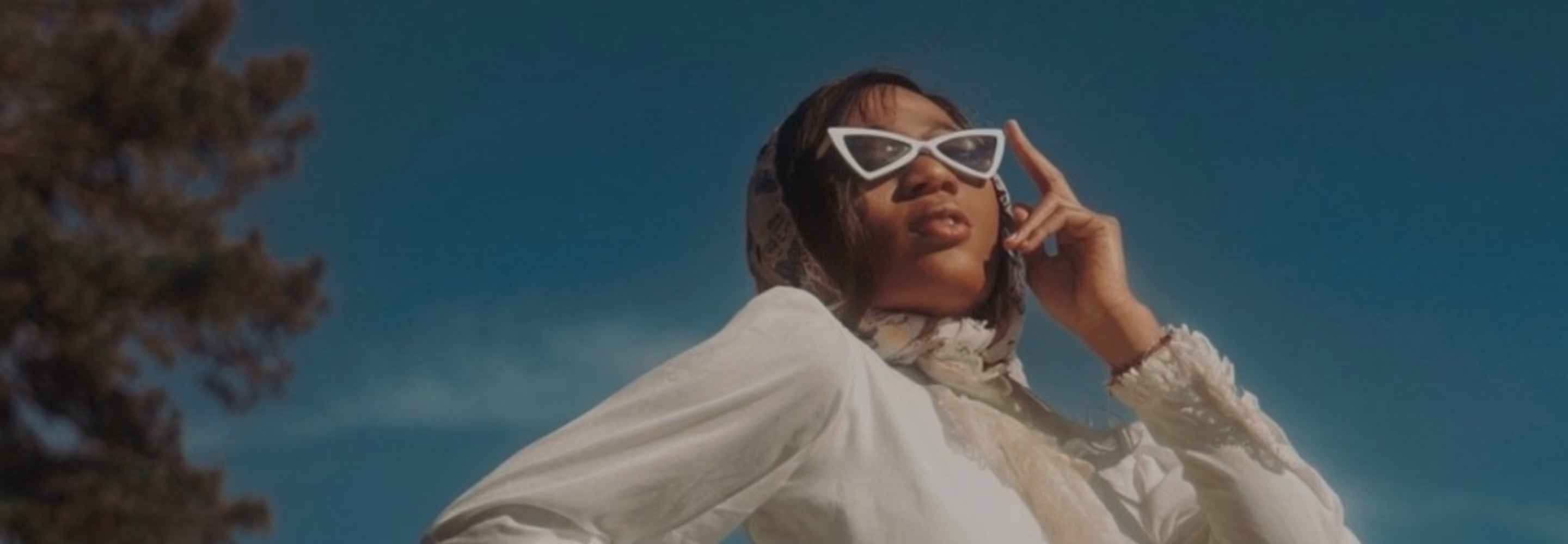 Person wearing white cat-eye sunglasses and a white outfit with a light scarf over their head posing outdoors against a clear blue sky with a tree on the left.