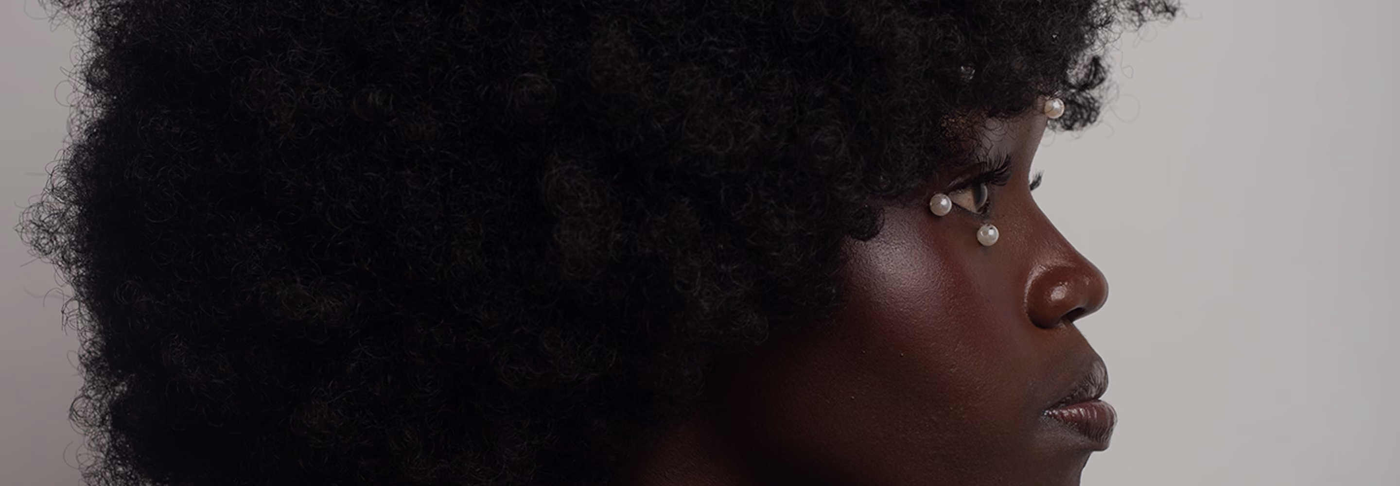 Close-up profile of a person with dark curly hair and dark skin, adorned with pearl-like beads near the eyes.