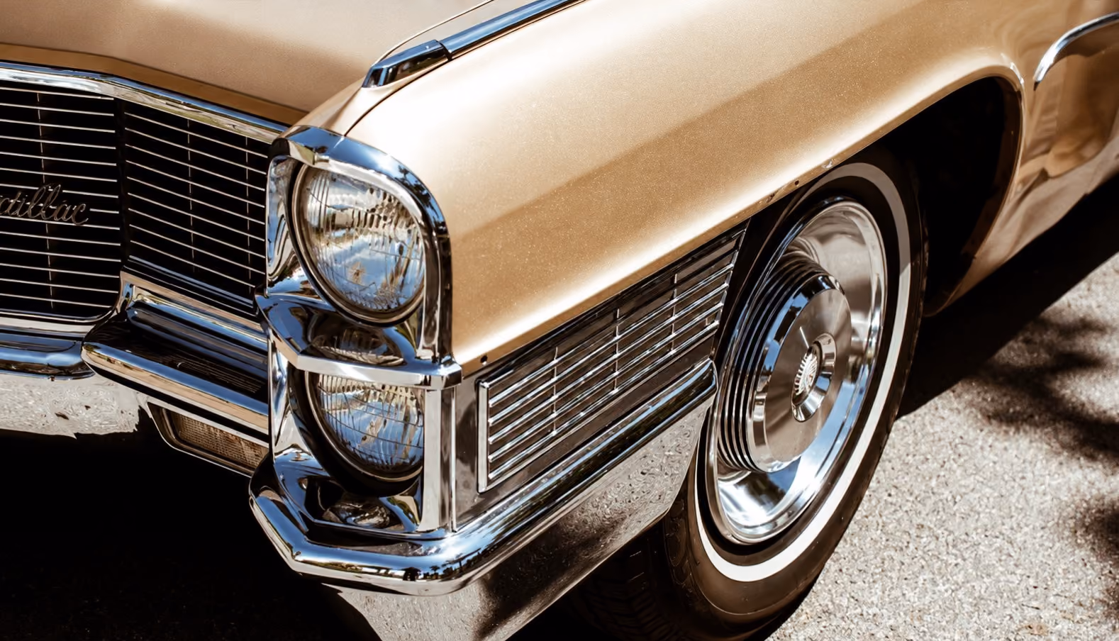 Vintage filter added to a gold Cadillac's front corner, showing its dual round headlights, chrome grille, bumper, and whitewall tire on a paved surface.