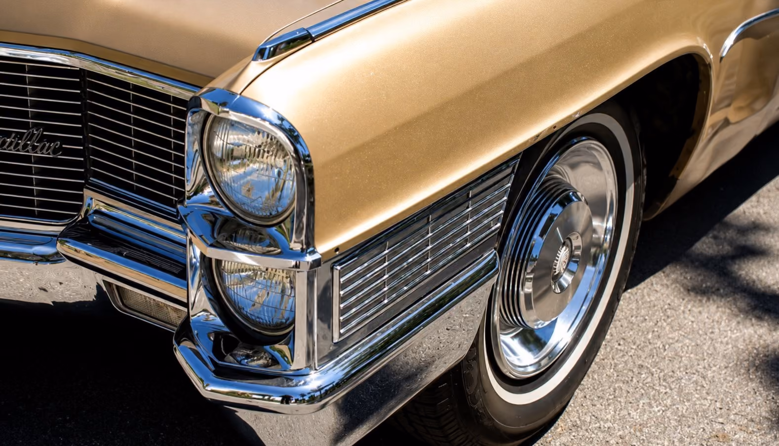 Original unedited image of a gold Cadillac's front corner, showing its dual round headlights, chrome grille, bumper, and whitewall tire on a paved surface.