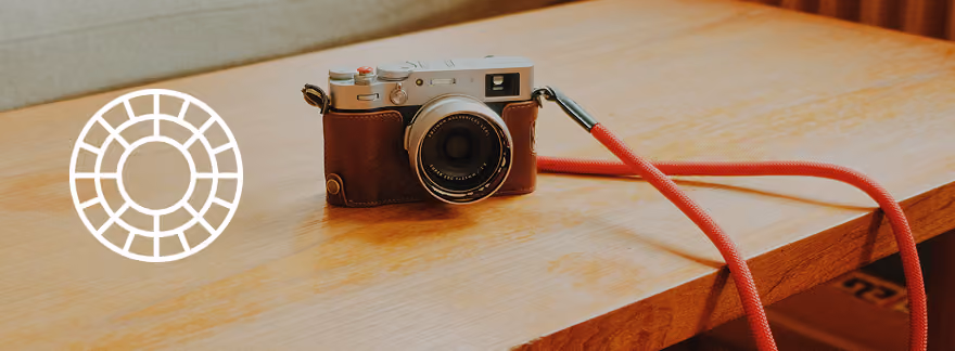 Vintage rangefinder camera with a brown leather case and a red strap resting on a wooden table with a beige wall in the background. VSCO logo overlay on left.