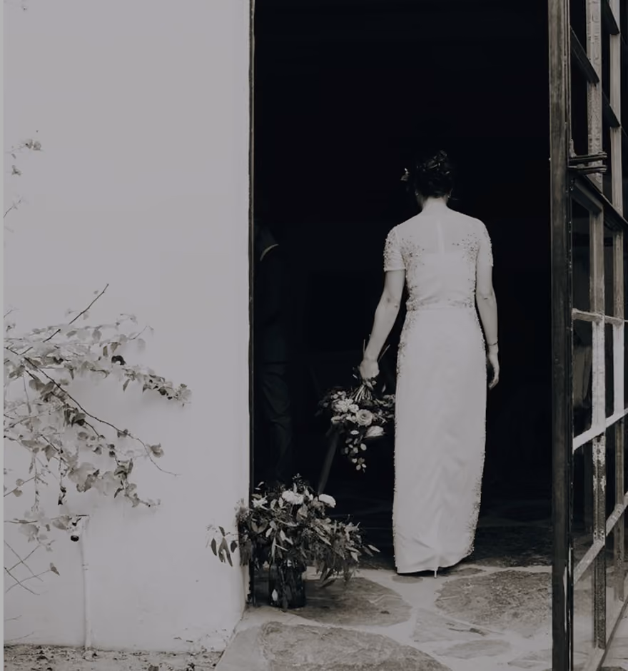 Black and white photo of a woman in a long, elegant dress holding a bouquet, walking into a dark doorway with plants beside the entrance.
