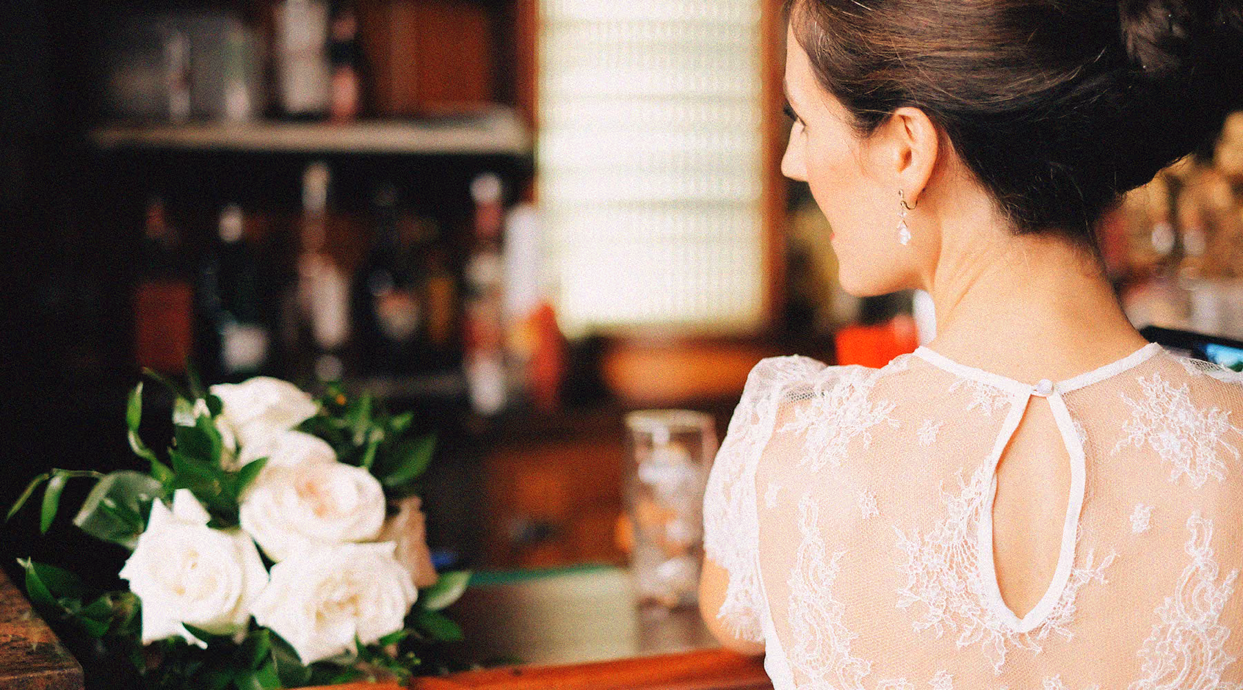 Close-up of a bride with her back to the camera, wearing a lace wedding dress and an updo hairstyle, with a bouquet of white roses on a wooden surface nearby.