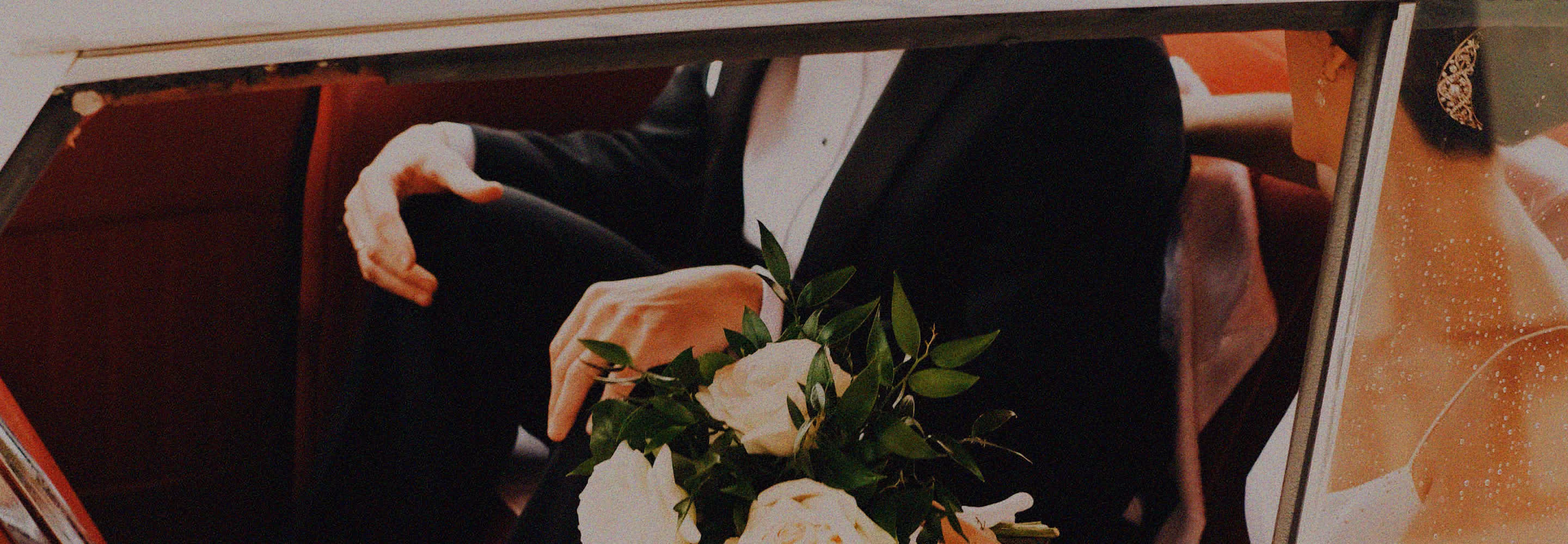 A person dressed in a black suit holding a bouquet of white flowers and greenery, visible through a car window.