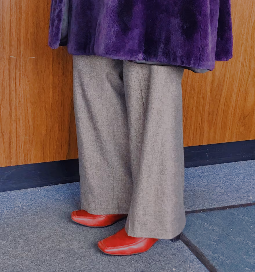 Person wearing wide-legged gray pants, bright red square-toed shoes, and a purple furry coat standing indoors on a carpeted floor next to a wooden wall.
