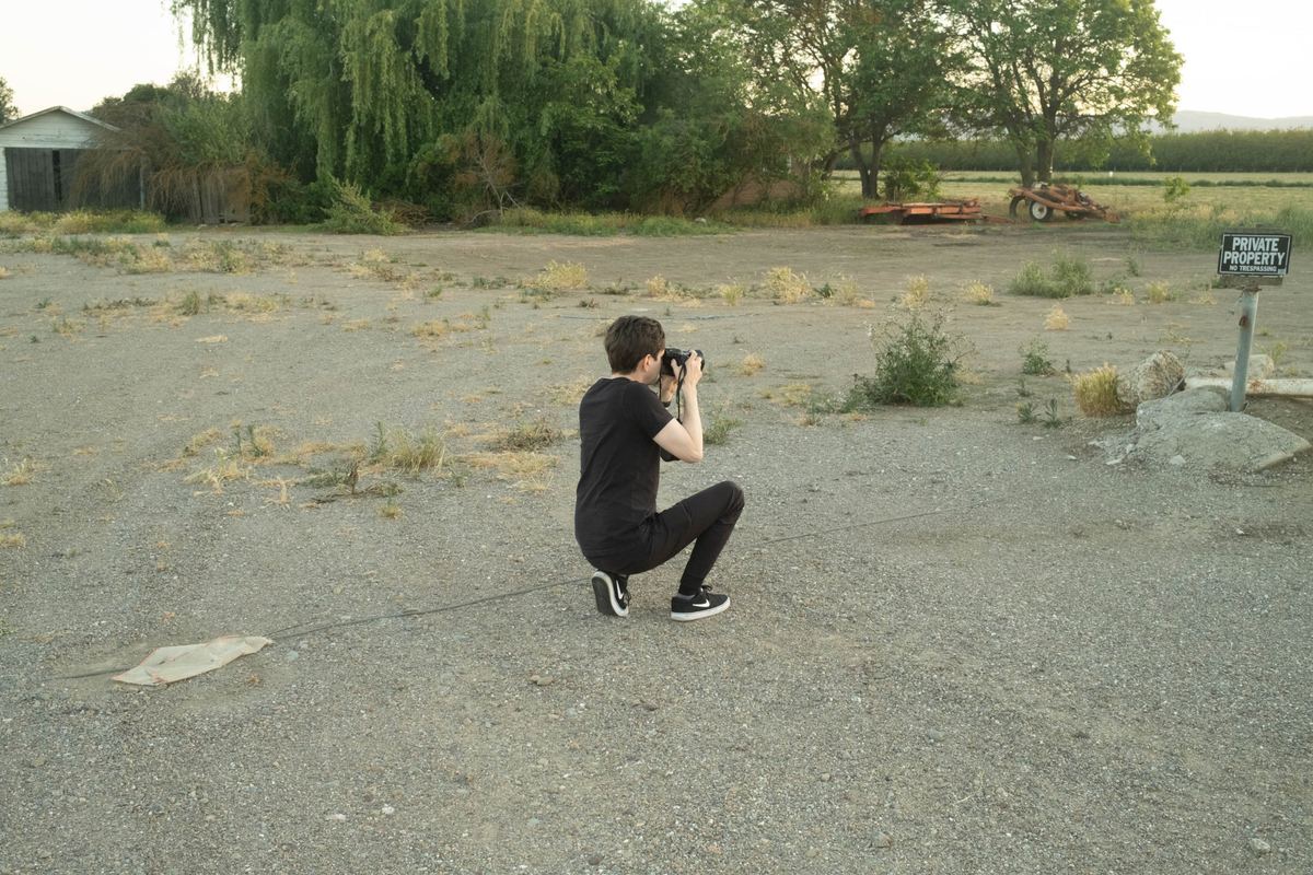 Photographer kneeling on a gravel lot to frame a shot with a DSLR, original unedited