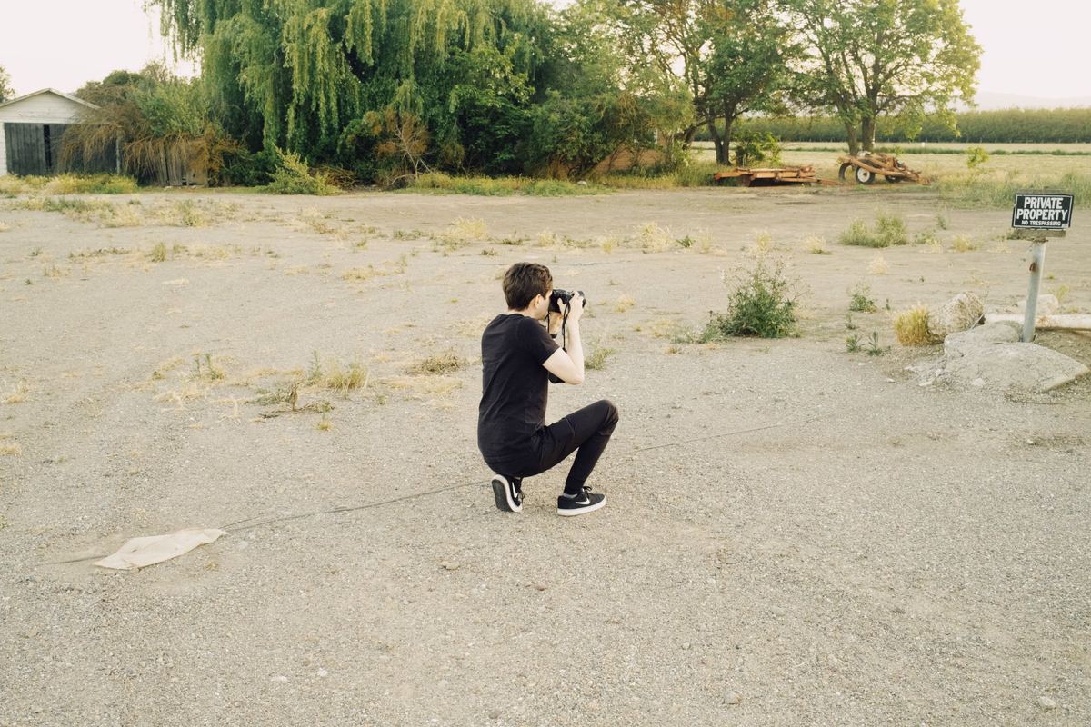 Photographer kneeling on a gravel lot to frame a shot with a DSLR, Kodak Portra 800 preset applied