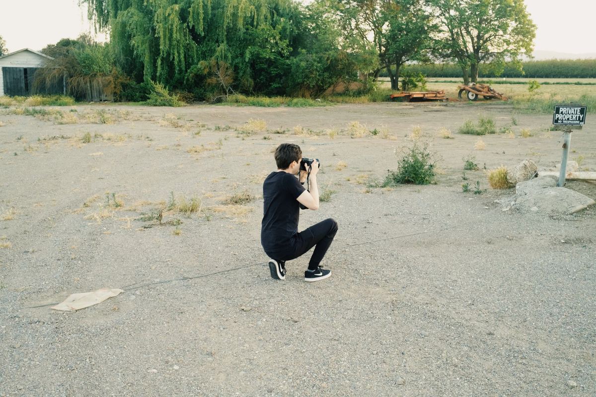 Photographer kneeling on a gravel lot to frame a shot with a DSLR, Kodak Ektar 100 preset applied