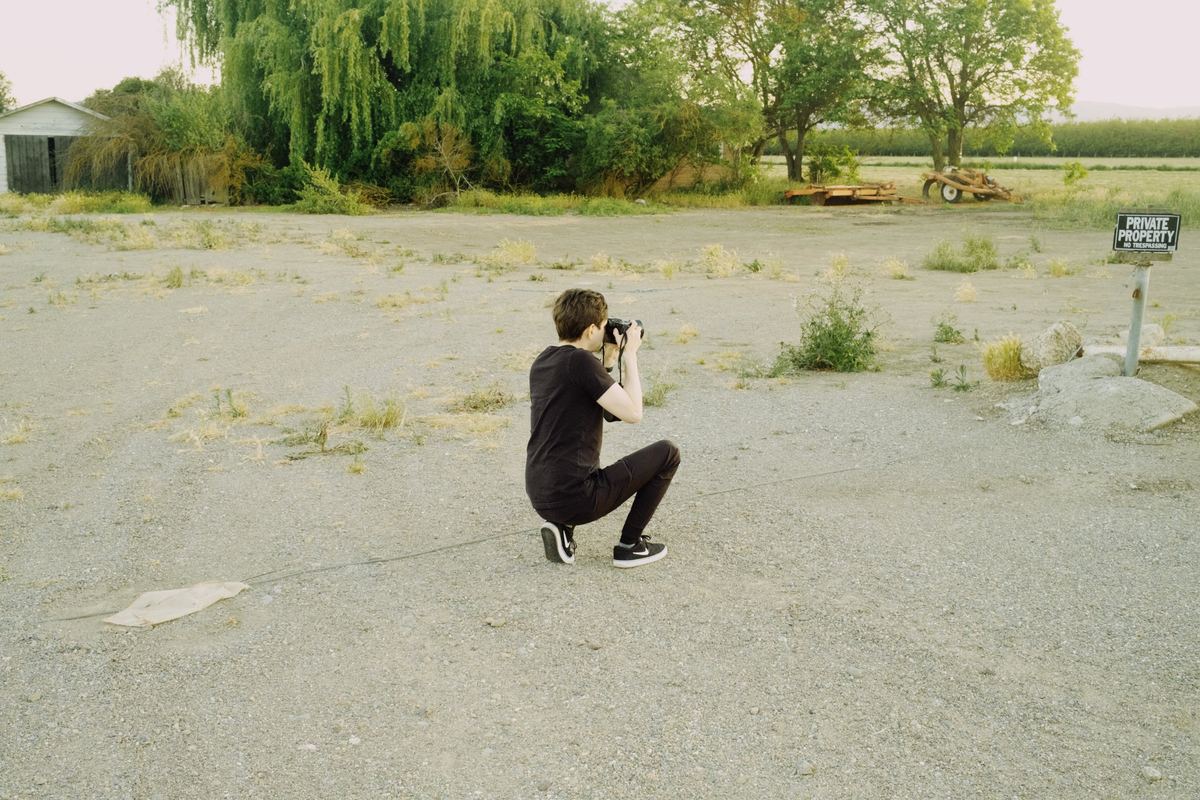 Photographer kneeling on a gravel lot to frame a shot with a DSLR, Kodak Gold 200 preset applied