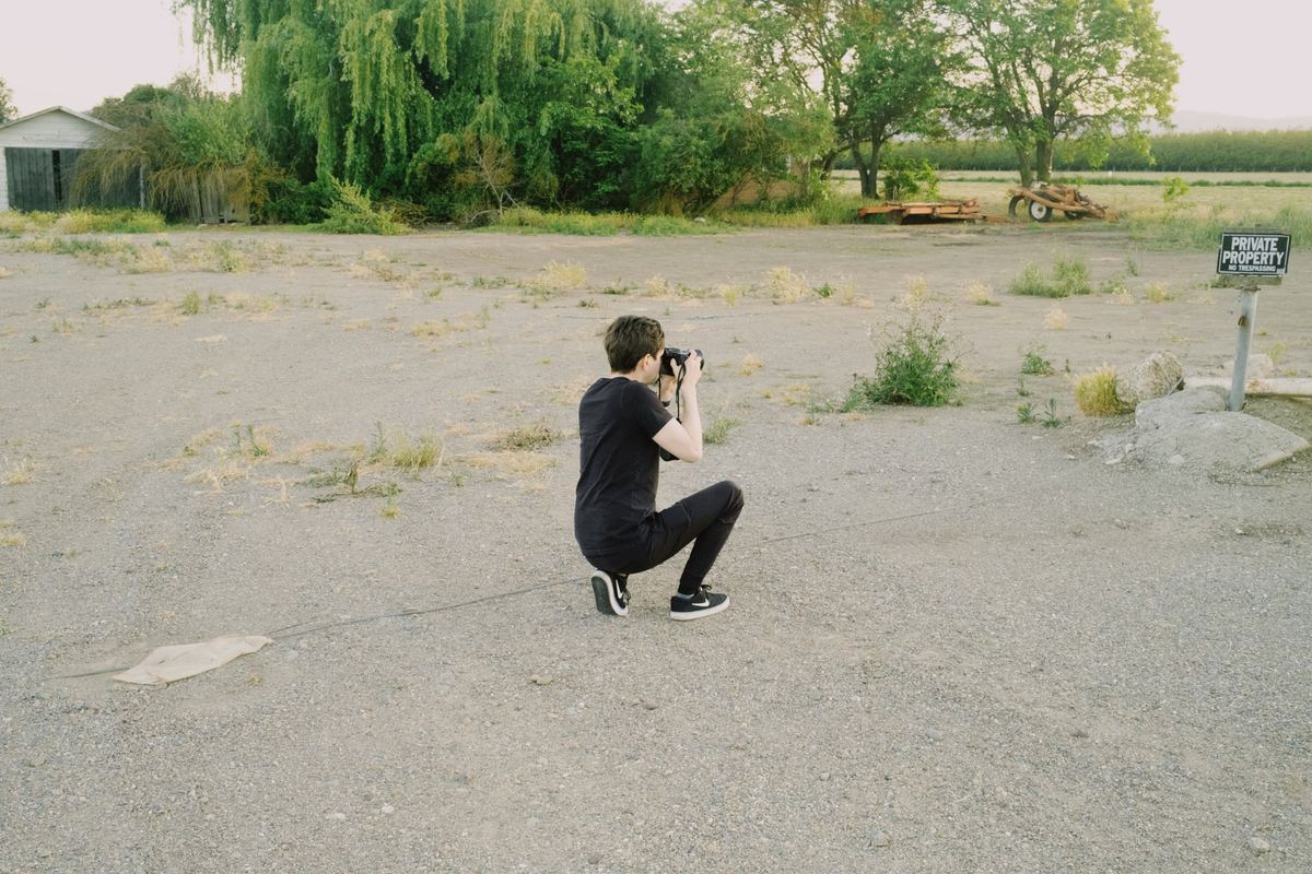 Photographer kneeling on a gravel lot to frame a shot with a DSLR, Fuji Pro 400H preset applied