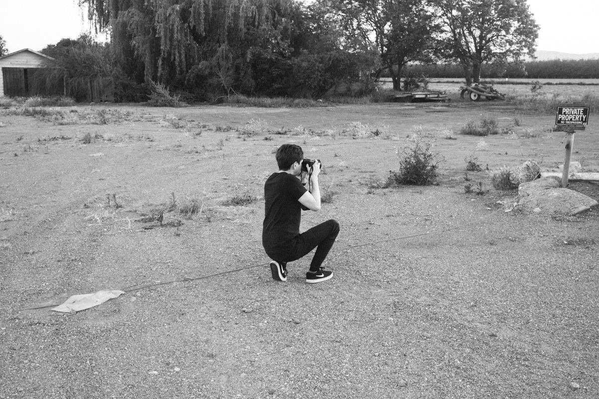 Photographer kneeling on a gravel lot to frame a shot with a DSLR, Ilford Delta 3200 preset applied