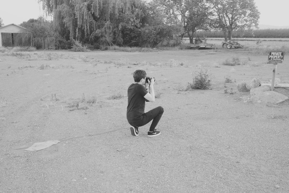 Photographer kneeling on a gravel lot to frame a shot with a DSLR, Ilford HP5 preset applied