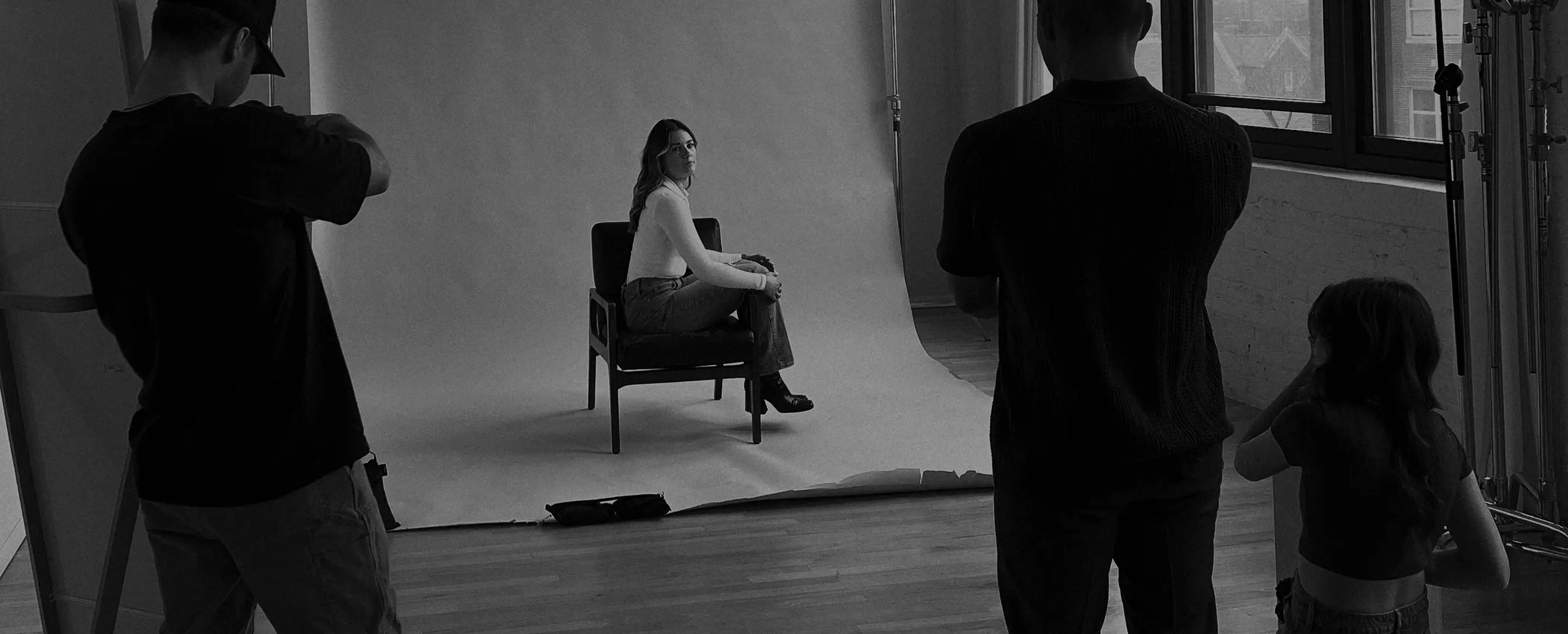 Black and white photo of photographers in a studio shooting a woman seated on a chair against a white backdrop.
