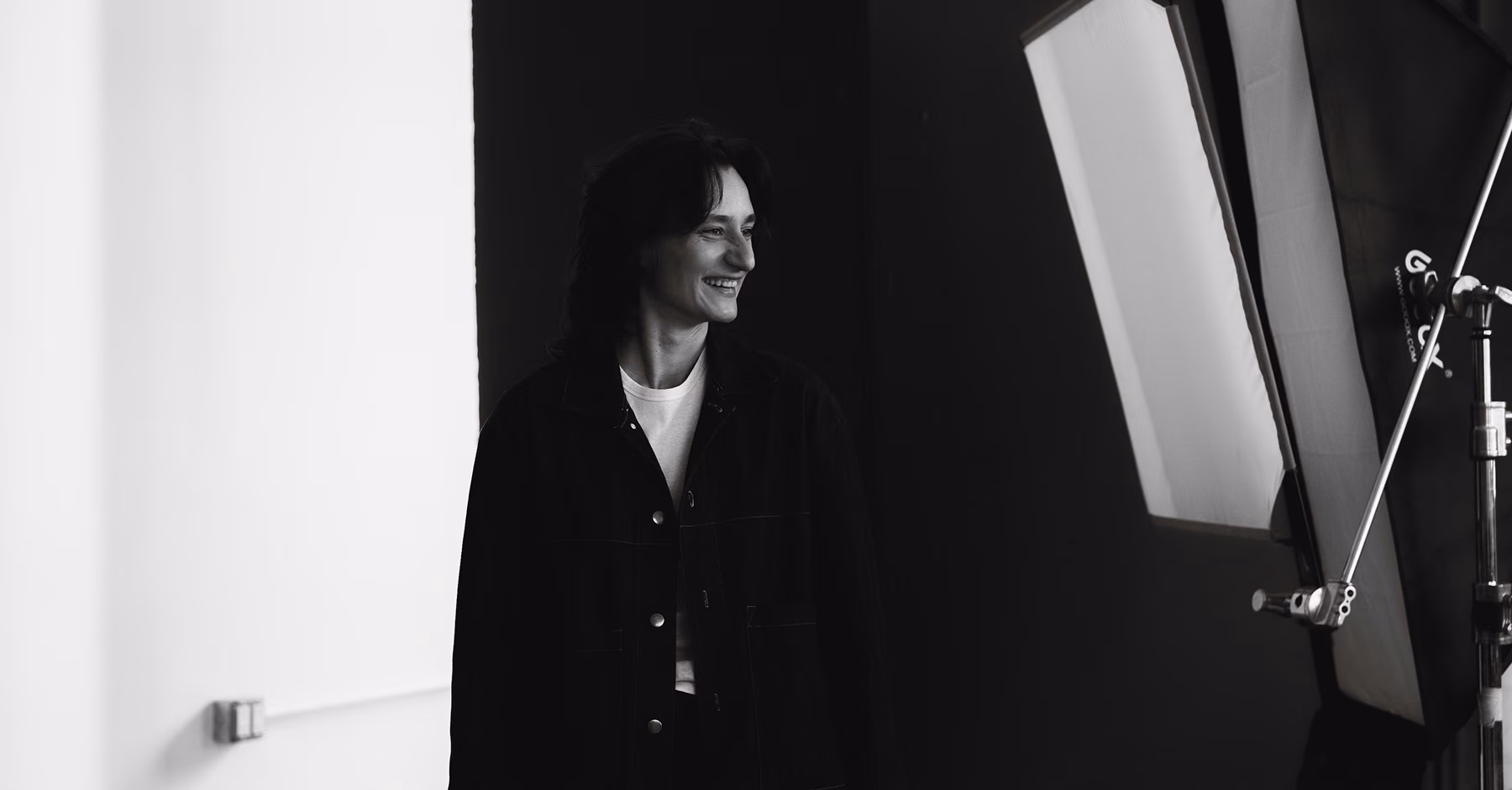 Smiling person in a dark jacket standing next to a softbox in a photo studio.