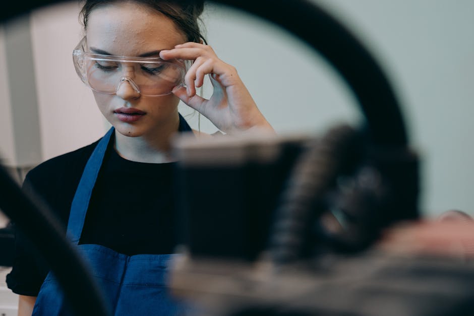 Young woman wearing safety glasses and apron in an industrial setting, focusing on her task.