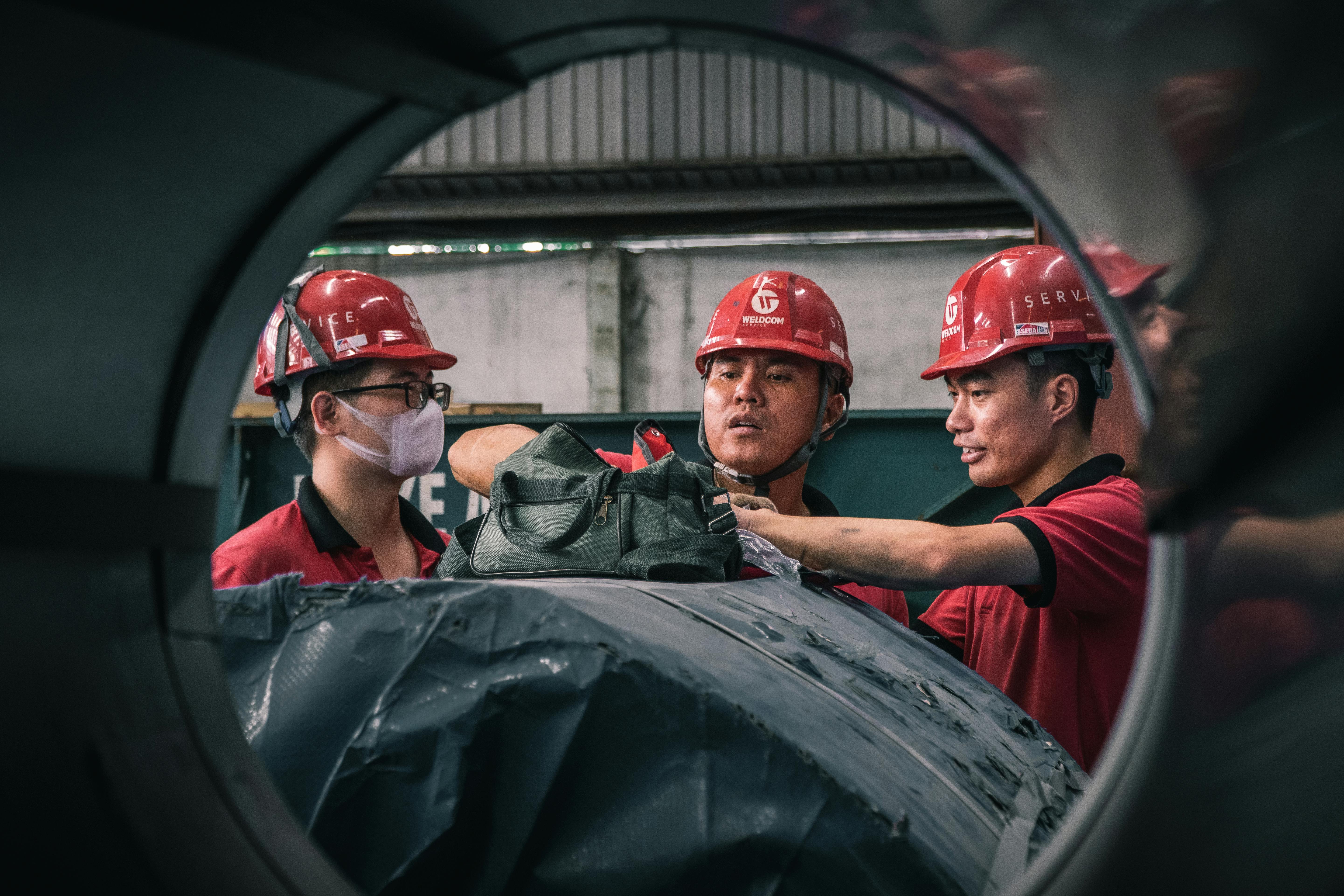 Three workers in safety helmets collaborate inside a factory setting, emphasizing teamwork and safety.