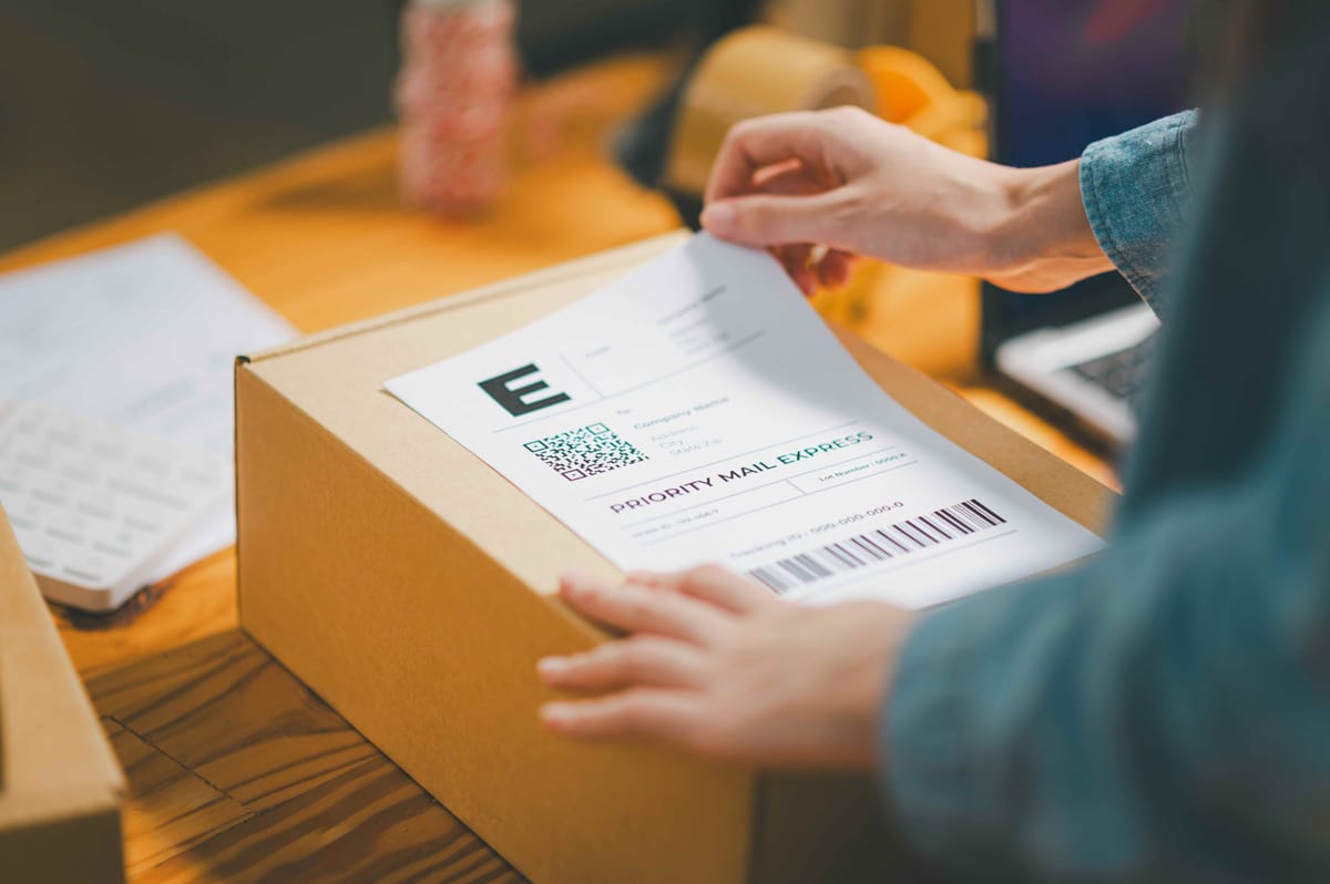 Person Attaching Shipping Label to Cardboard Box for Priority Mail Express on Wooden Desk with Office Supplies in Background