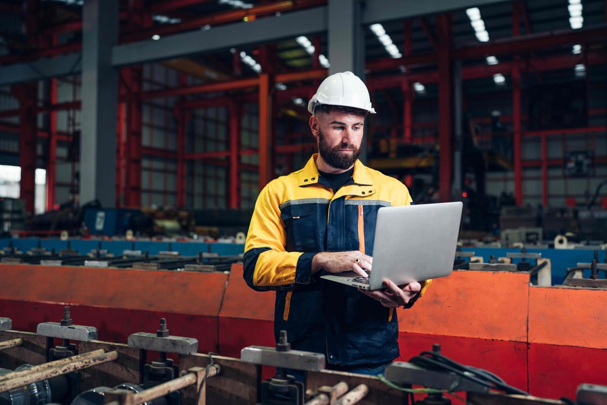 Industrial Engineer in Hard Hat and Reflective Jacket Using Laptop in a Modern Factory Setting with Machinery and Metal Structures
