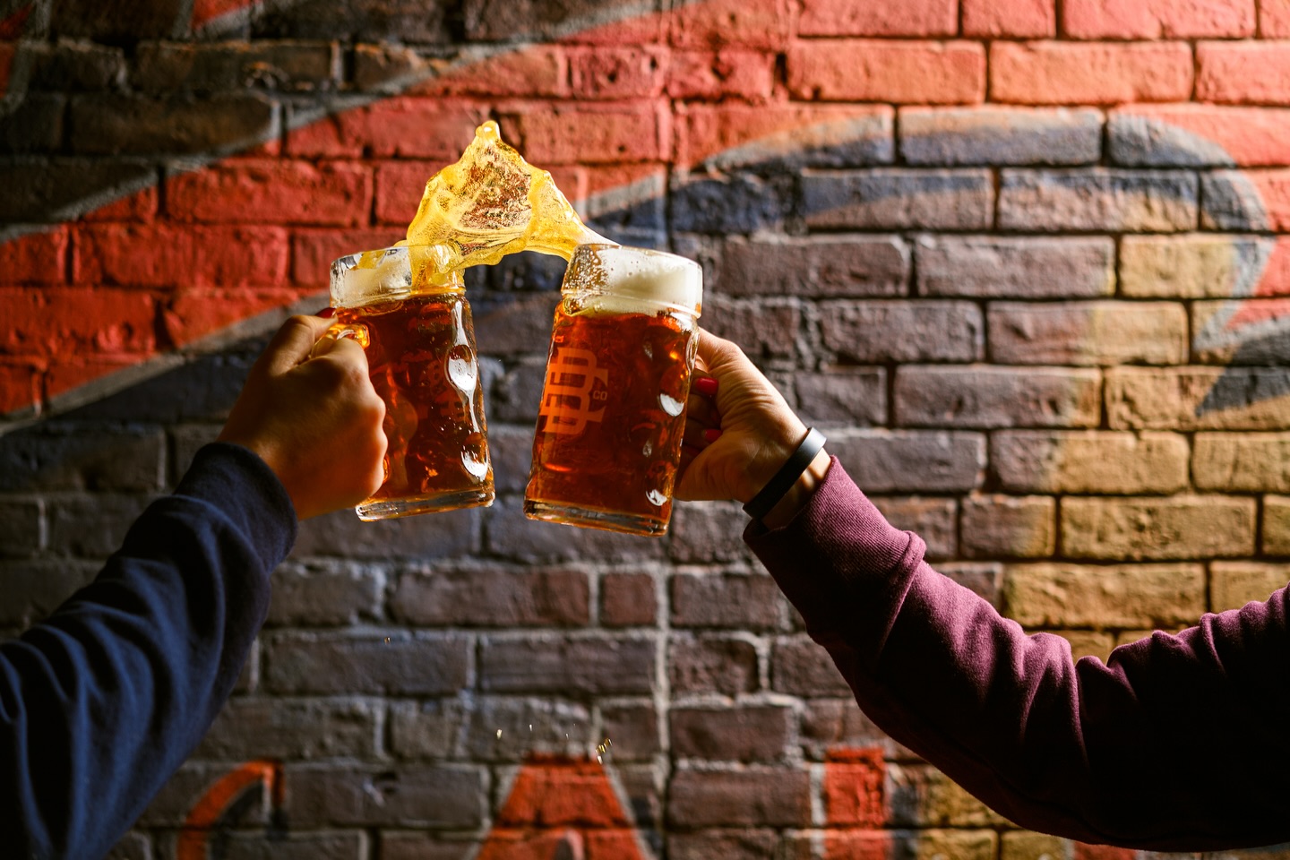 Two people clinking beer mugs with foam splashing at Elicit Brewing, against a brick wall background.