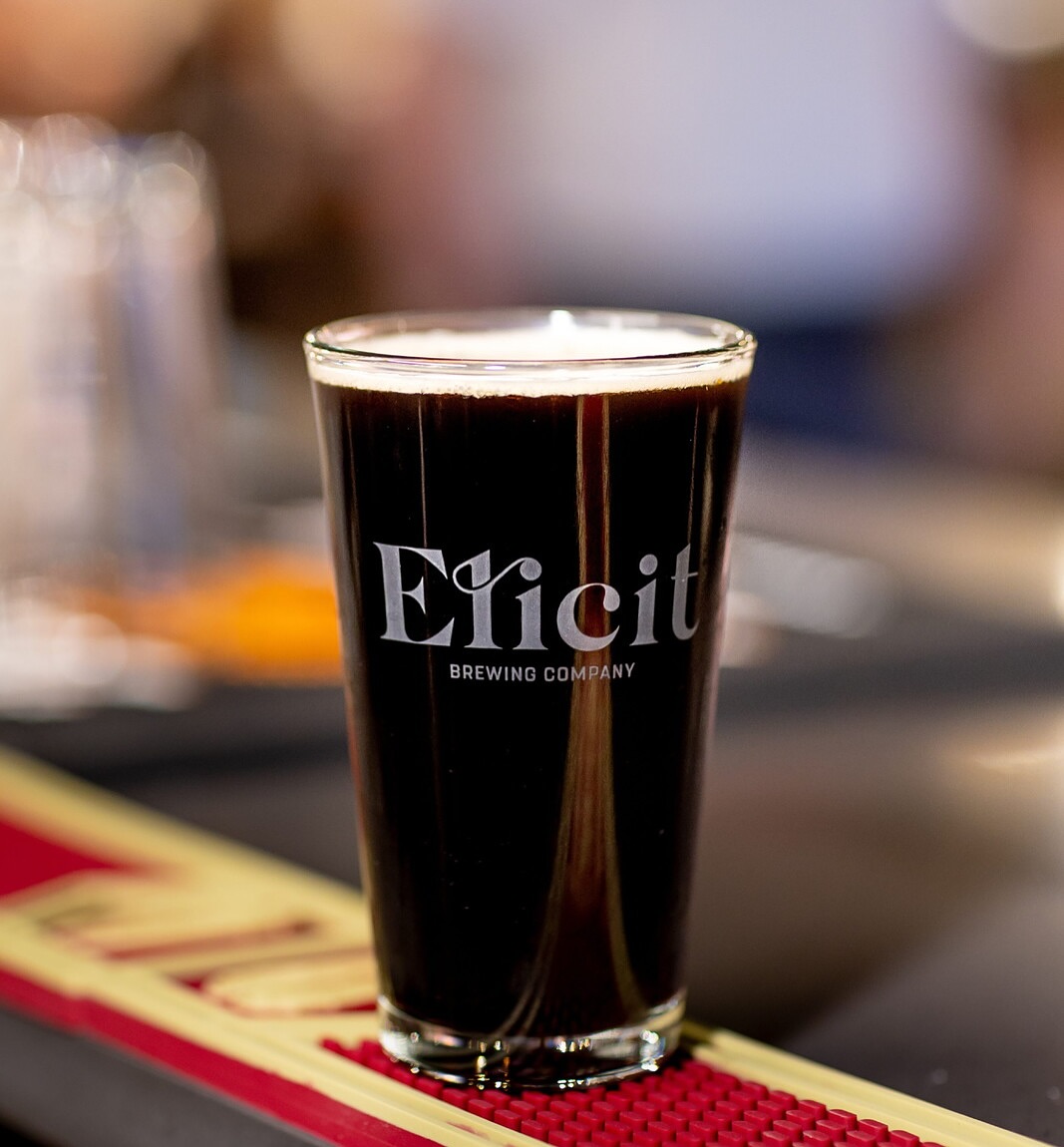 An image of a Dark stout beer in an Elicit Brewing Company glass on a wooden table.