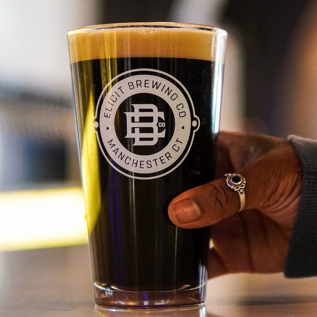  An image of a hand holding a pint of dark beer in an Elicit Brewing Manchester, CT glass.