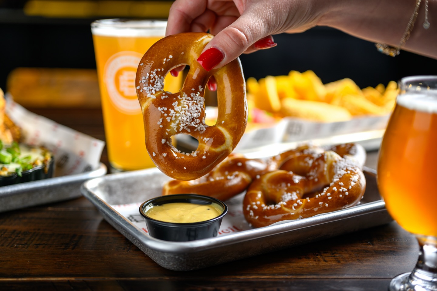 A hand holding a salted soft pretzel over a tray of pretzels, dip, and beer at Elicit Brewing.